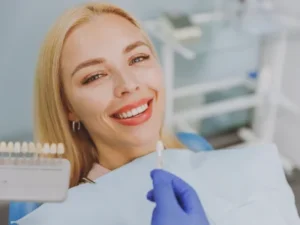 A patient smiling during a cosmetic dentistry consultation while a dentist uses a shade guide to select dental veneers.